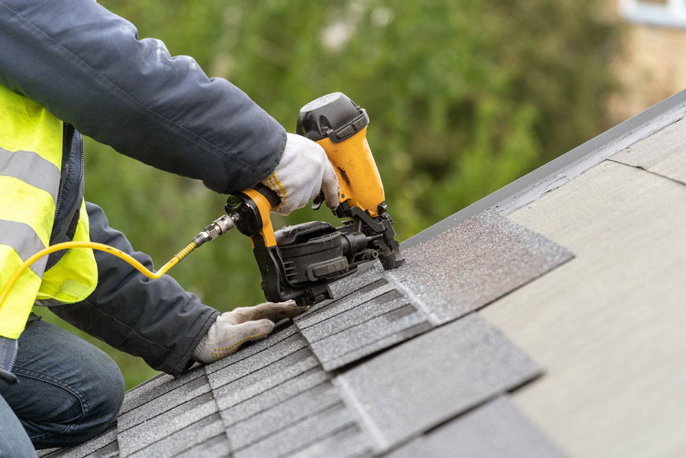 Roofer using a nail gun to attach a gray asphalt roofing shingle to a roof